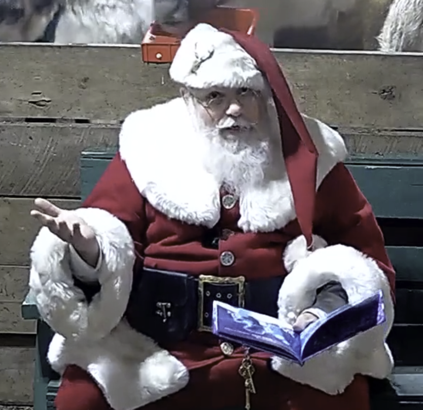 Santa sitting in green chair holding book open in left hand, gesturing out and upward with his left